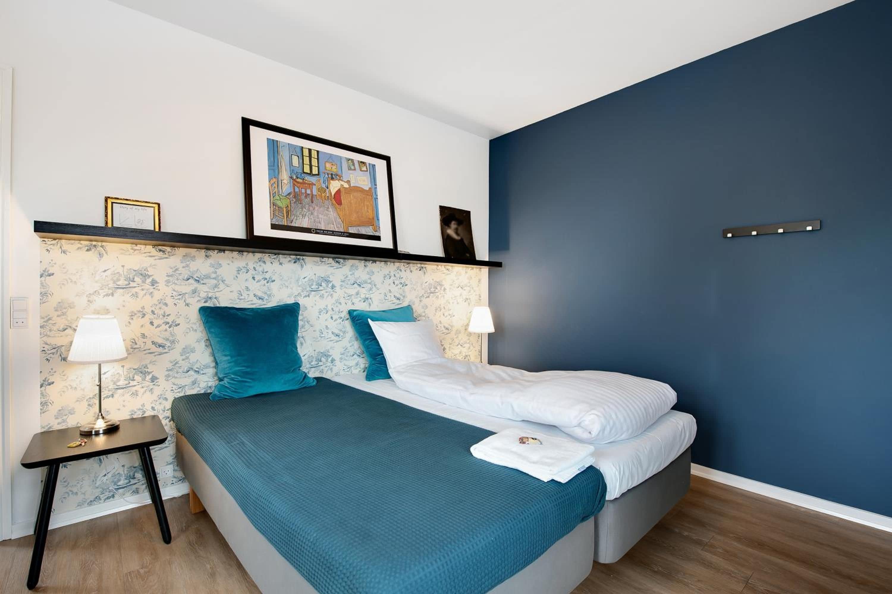 A modern bedroom with a bed, two bedside tables, a nightstand, a framed picture, and a wall-mounted light fixture.