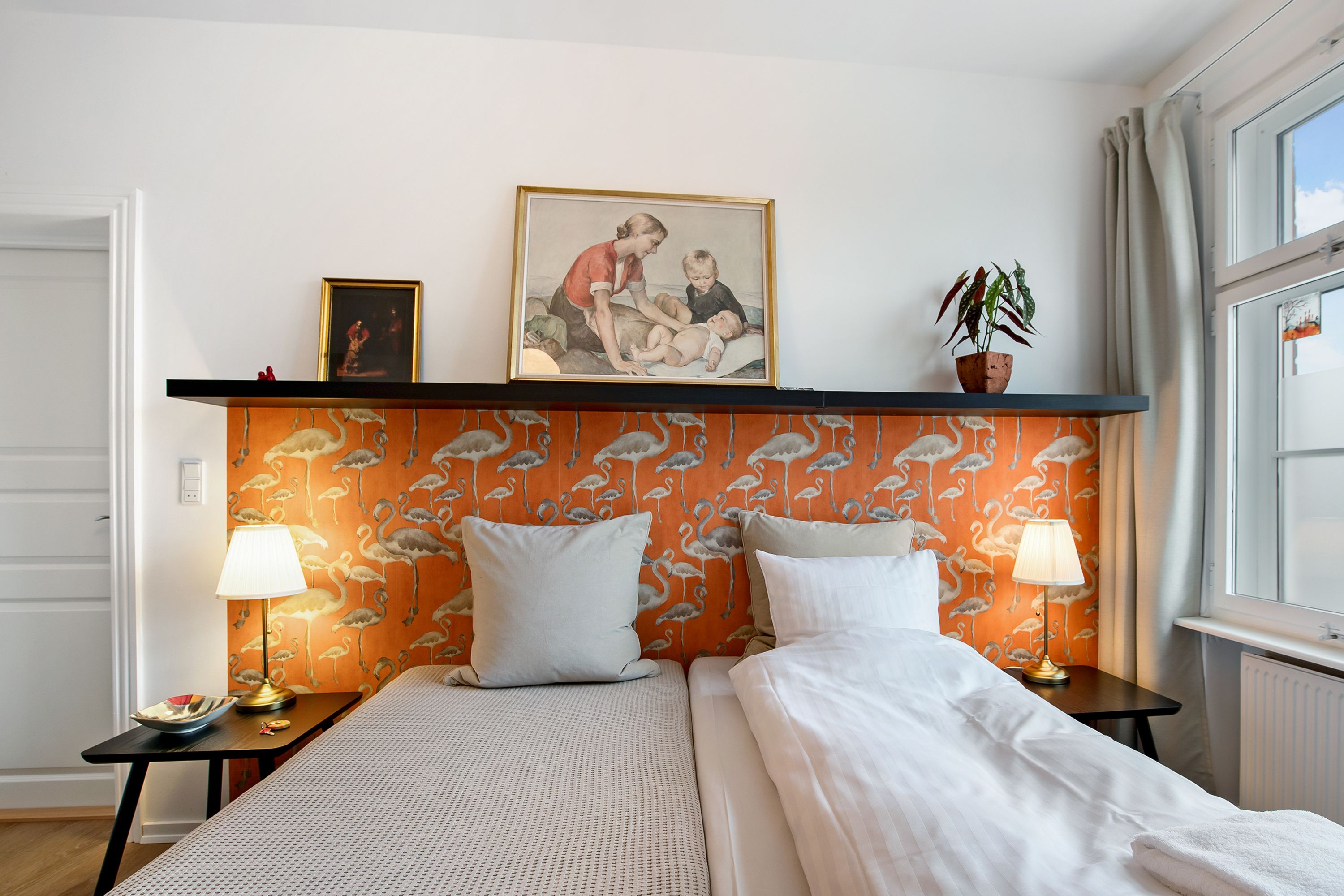 A hotel room with a bed, two bedside tables, a nightstand, a framed picture, a potted plant, and a window with curtains.