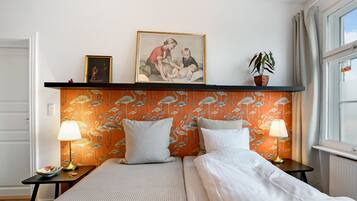 A hotel room with a bed, two bedside tables, a nightstand, a framed picture, a potted plant, and a window with curtains.