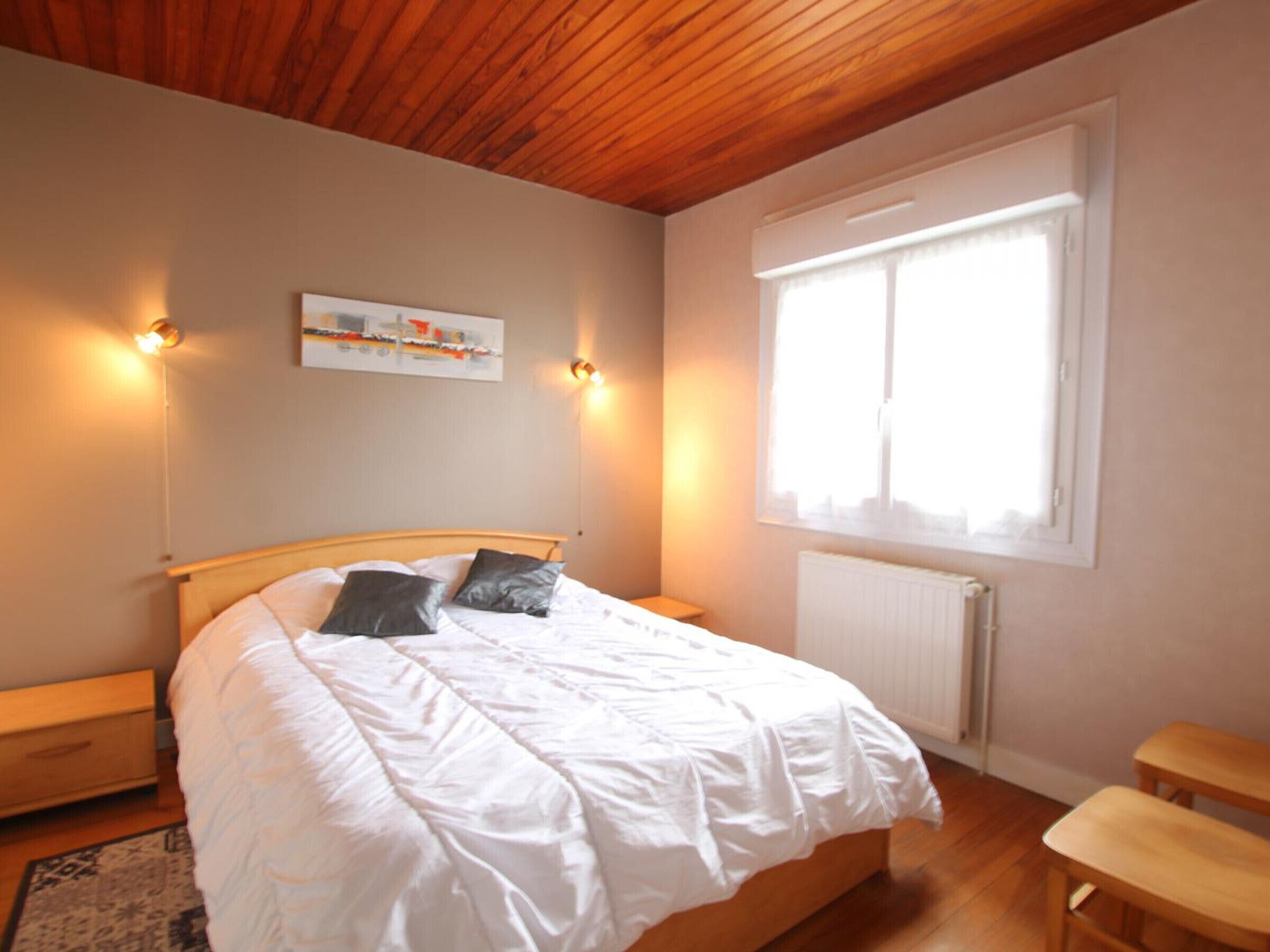 A bedroom with a wooden ceiling, a bed with white bedding, two bedside lamps, a radiator, and a window with white curtains.