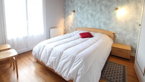 Une chambre à coucher avec un lit en bois, du linge de lit blanc, un oreiller rouge, une table de chevet, une petite table et un tapis à motifs.