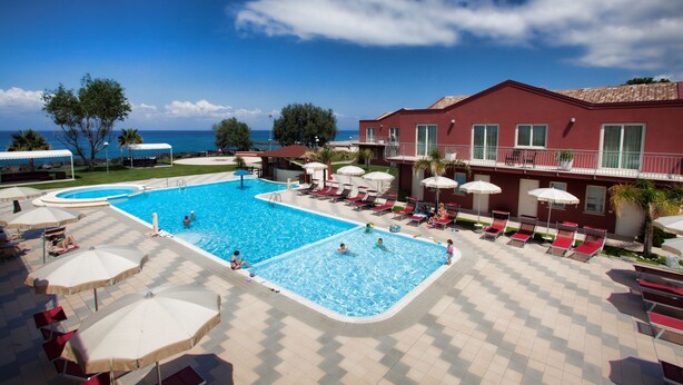 A swimming pool area with lounge chairs and umbrellas, a red building with balconies, and a view of the sea. - Hotel Valeria del Mar (Belvedere Marittimo)