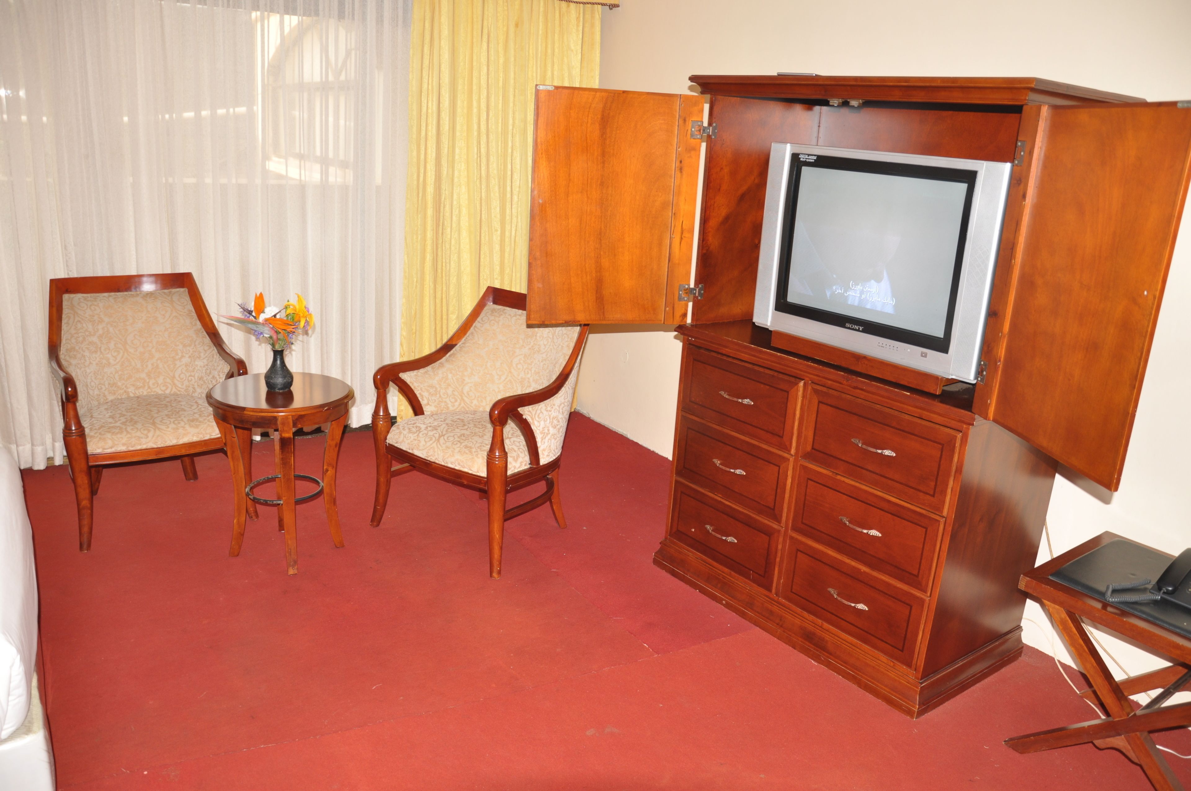 A room with two wooden armchairs, a small round table with a vase of flowers, a wooden cabinet with a television, and a red carpet.