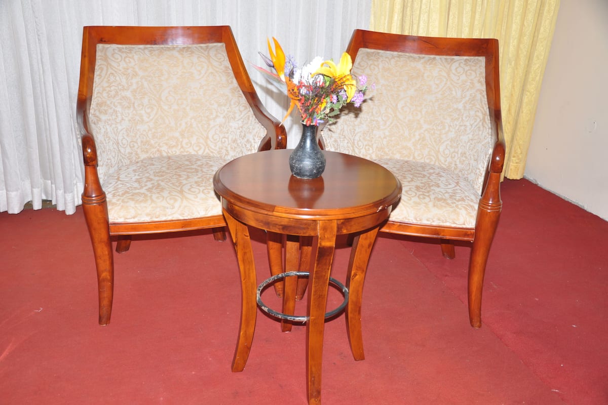 Duas cadeiras de madeira com assentos e encostos acolchoados, uma pequena mesa redonda de madeira com um vaso de flores, e um chão coberto por um tapete vermelho.