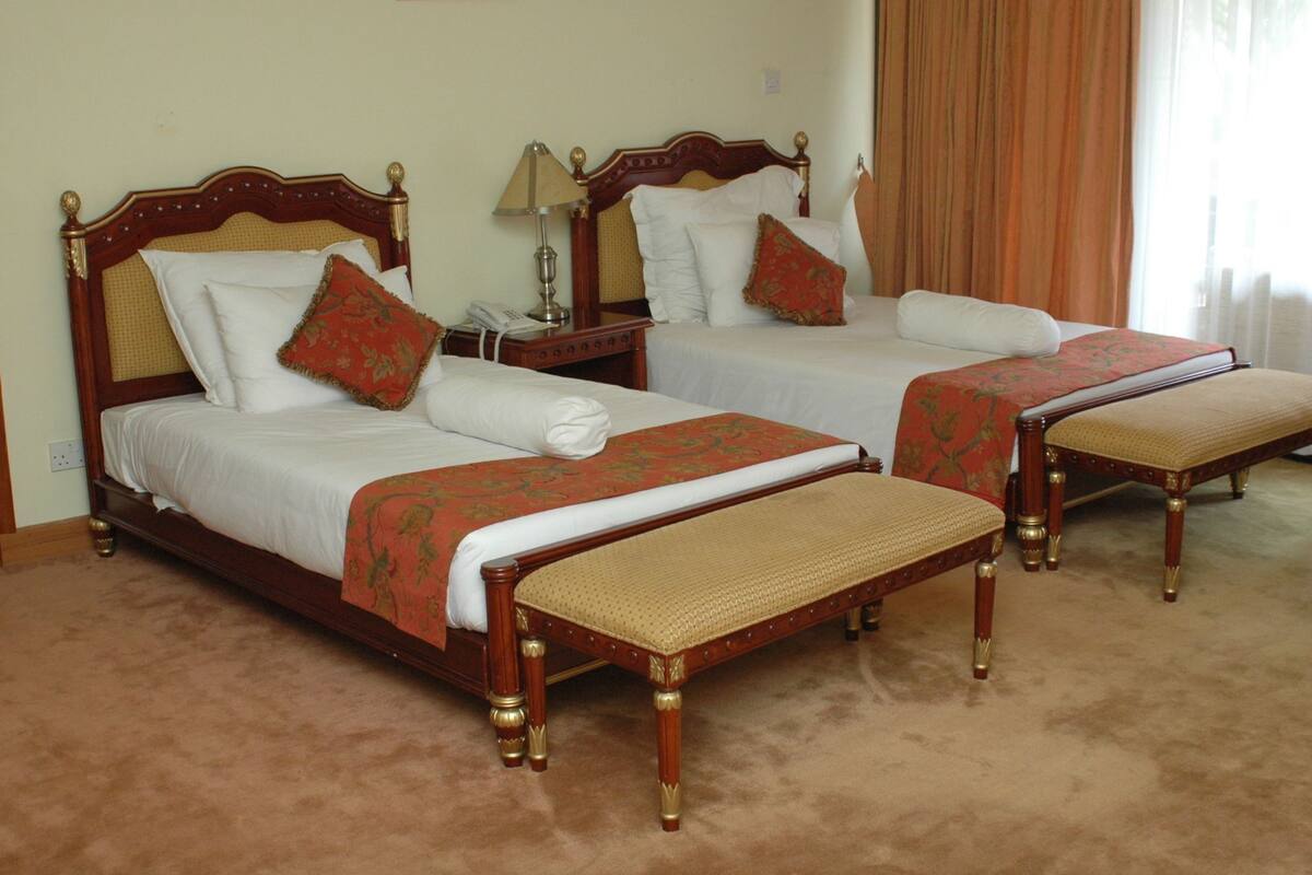 Quarto de hotel com duas camas de madeira, cada uma com cabeceira, mesas de cabeceira e um banco.