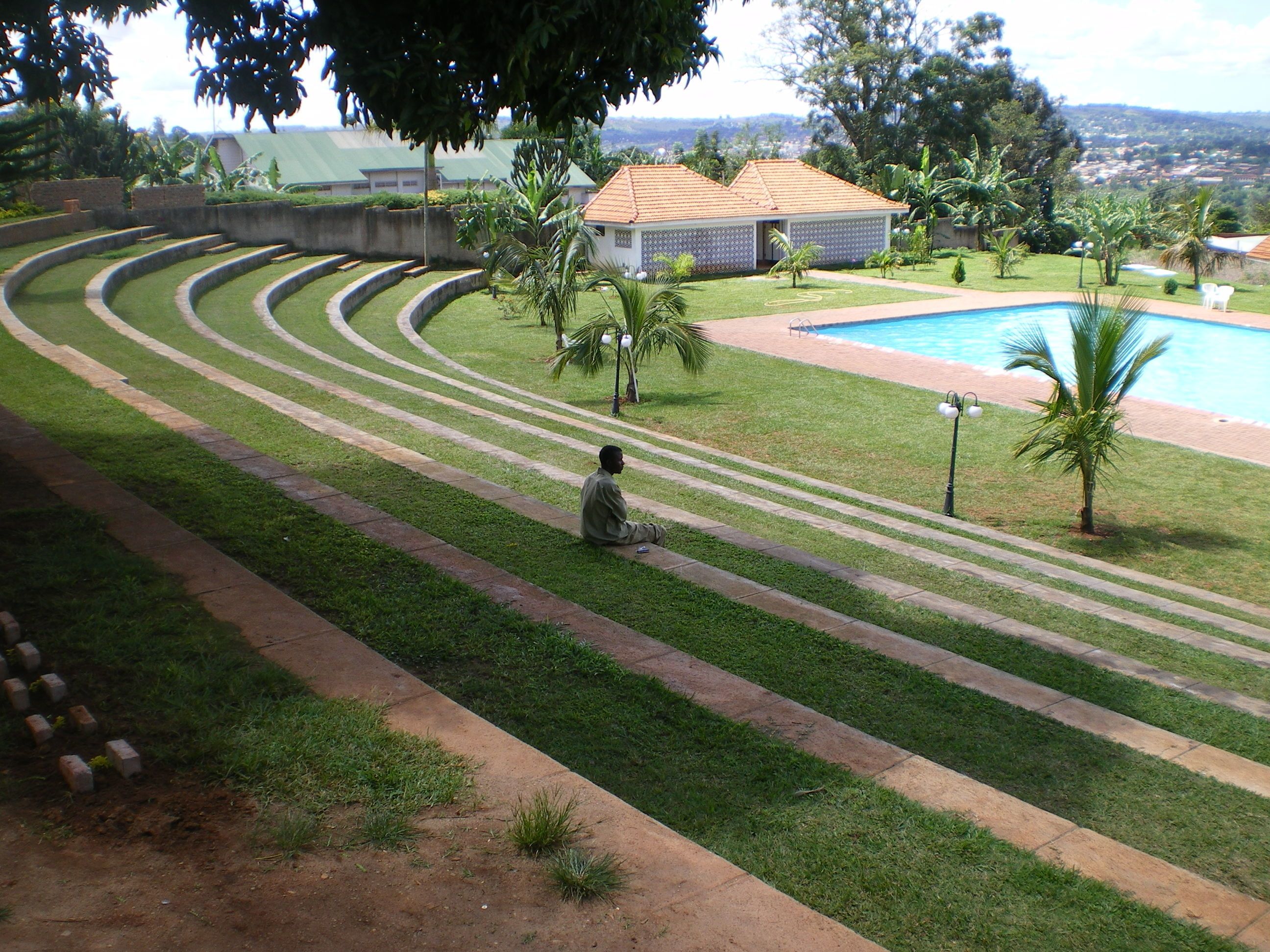 Un espace piscine avec un chemin sinueux, un petit bâtiment et une personne assise sur l’herbe.
