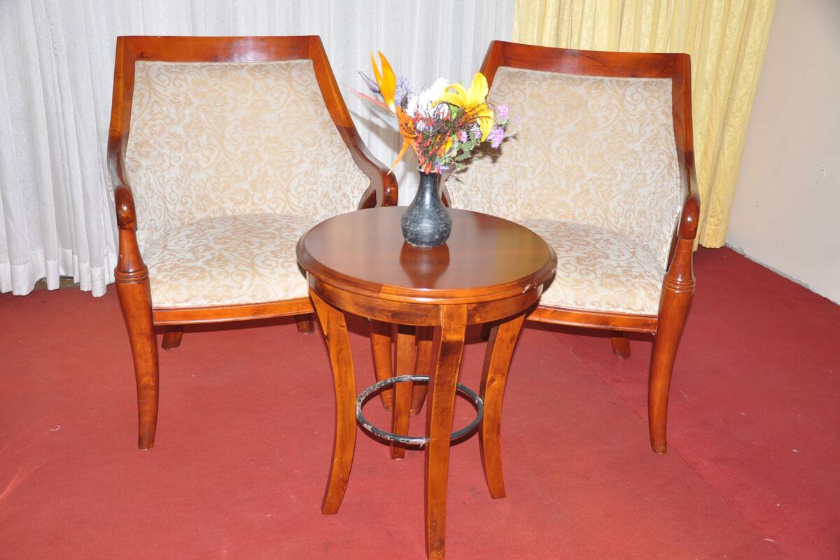 Duas cadeiras de madeira com assentos e encostos acolchoados, uma mesa de madeira redonda com um vaso de flores e um chão coberto por um tapete vermelho.