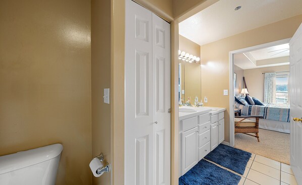 Une salle de bain avec des toilettes, un lavabo et un miroir avec éclairage. Il y a un tapis bleu sur le sol et une porte qui donne sur une chambre.