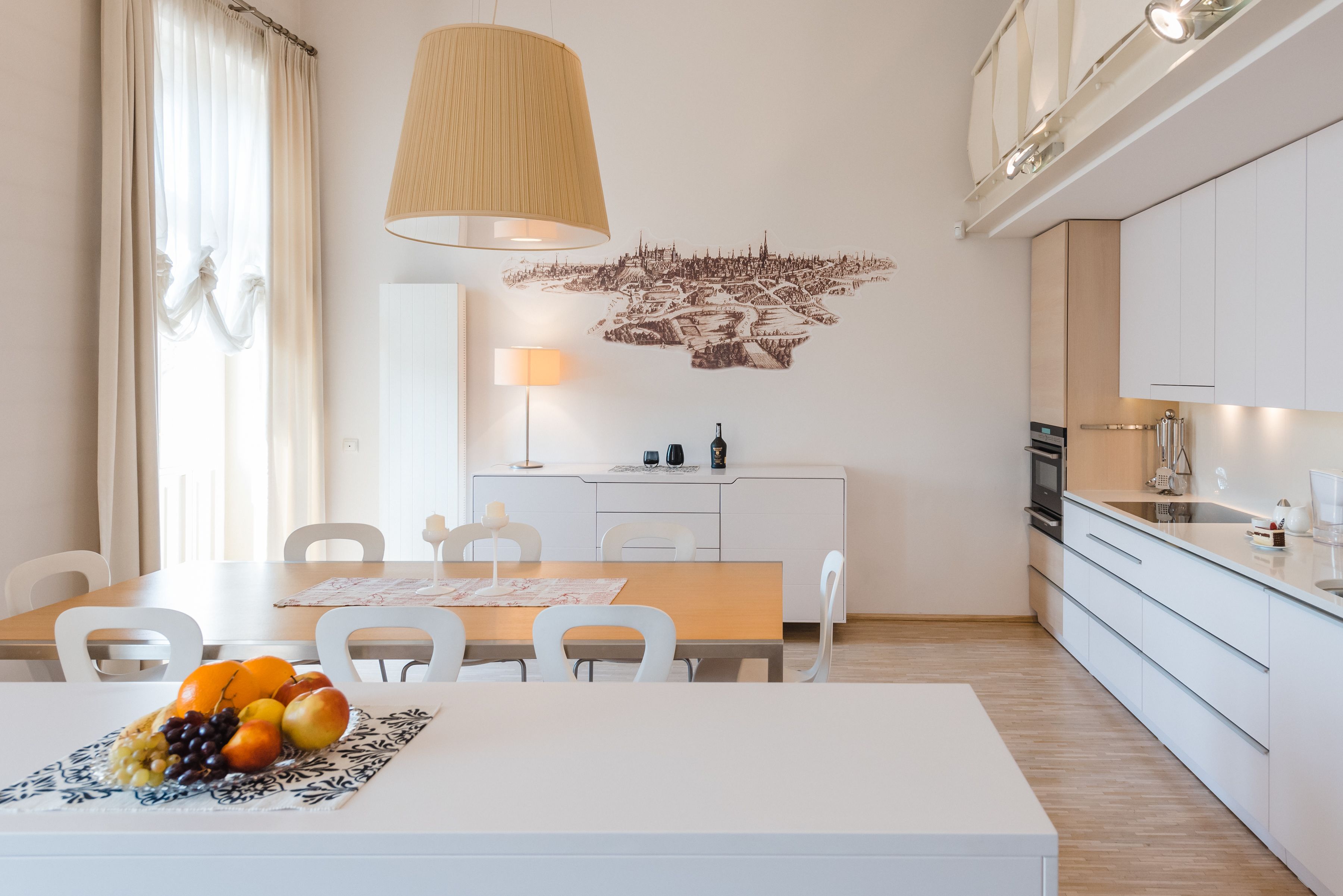 Une cuisine moderne avec des meubles blancs, une table à manger avec des chaises et un panier de fruits.