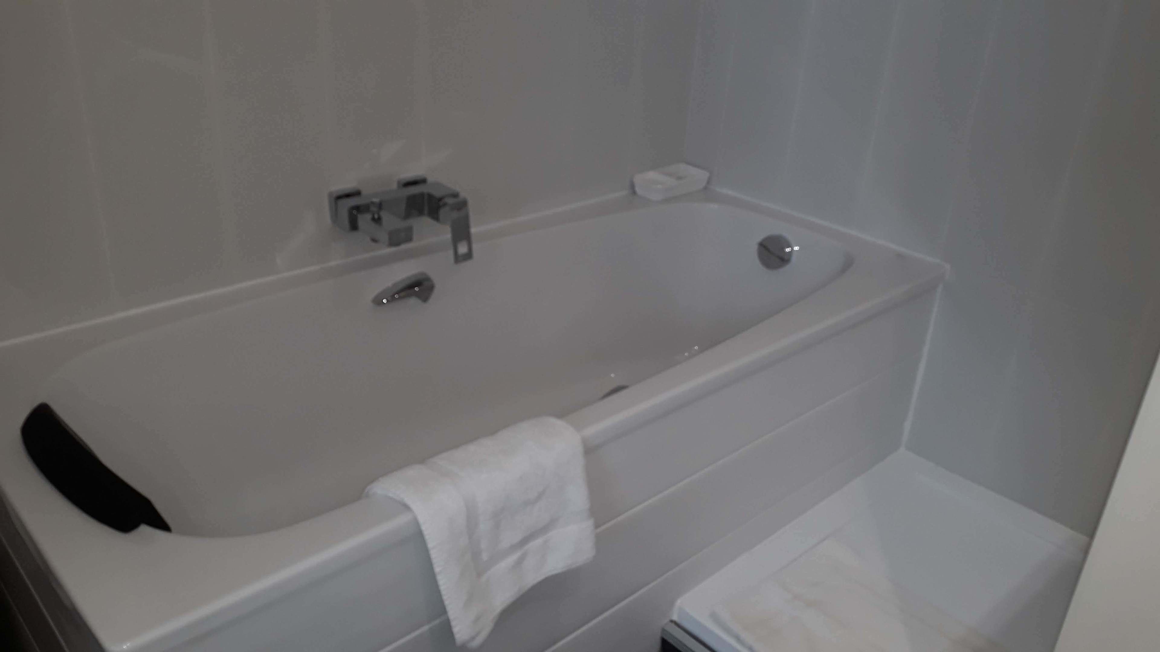 A modern bathroom with a white bathtub, a handheld showerhead, and a small white towel on the floor.