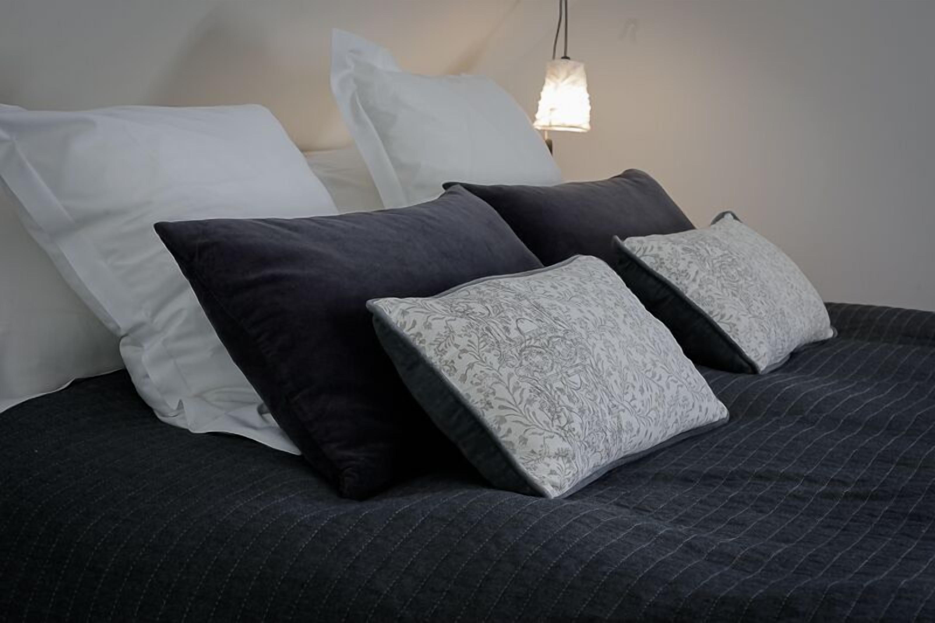 Una cama con una colcha acolchada oscura y cuatro almohadas en diferentes tonos de gris y blanco.