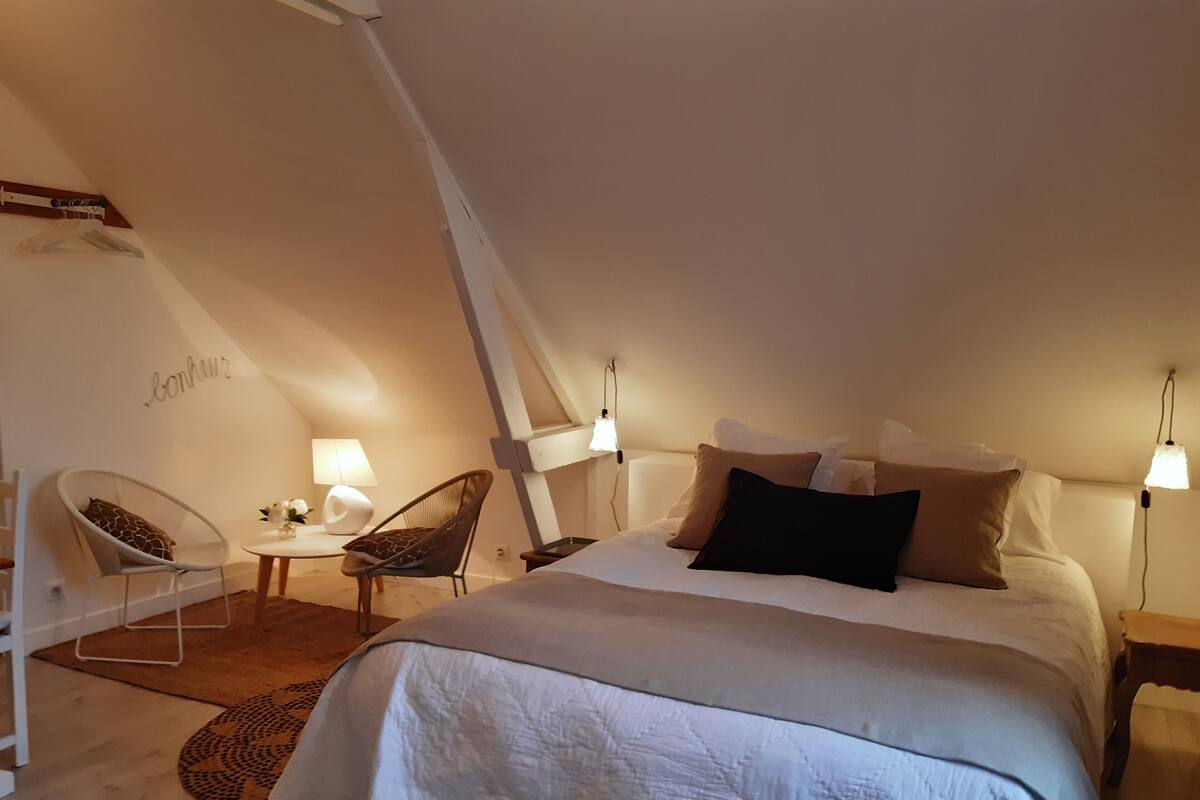 A cozy, well-lit attic bedroom with a large bed, two chairs, a small table, and a 'bonjour' sign on the wall.
