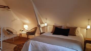 Une chambre mansardée agréable et bien éclairée, avec un grand lit, deux fauteuils, une petite table et un panneau indiquant « bonjour » sur le mur.