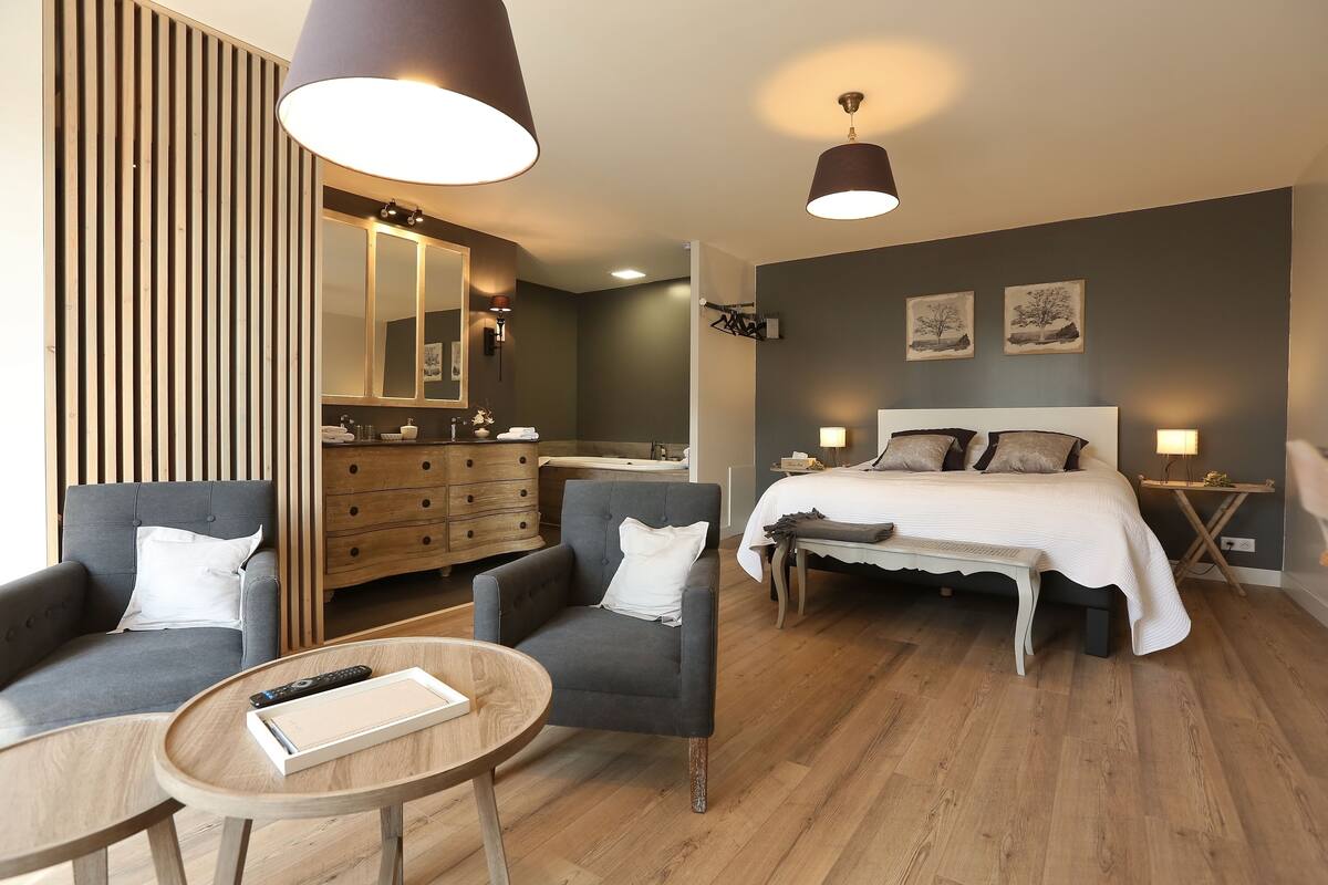 A modern hotel room with a sofa, armchair, round table, and a bed with a white bedspread.