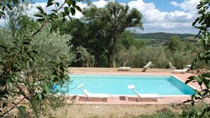 A rectangular pool with clear blue water, surrounded by a brick deck. There are four lounge chairs aligned along the pool's edge. The pool is set in a natural landscape with trees and hills in the background. - Agriturismo Raggio di Sole (Montepulciano)