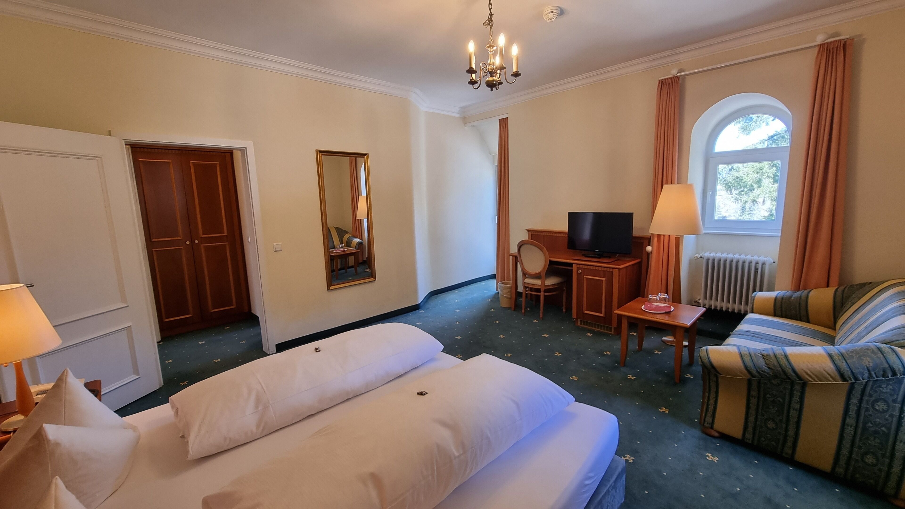 A hotel room with a bed, a sofa, a desk with a TV, a mirror, and a window with curtains.