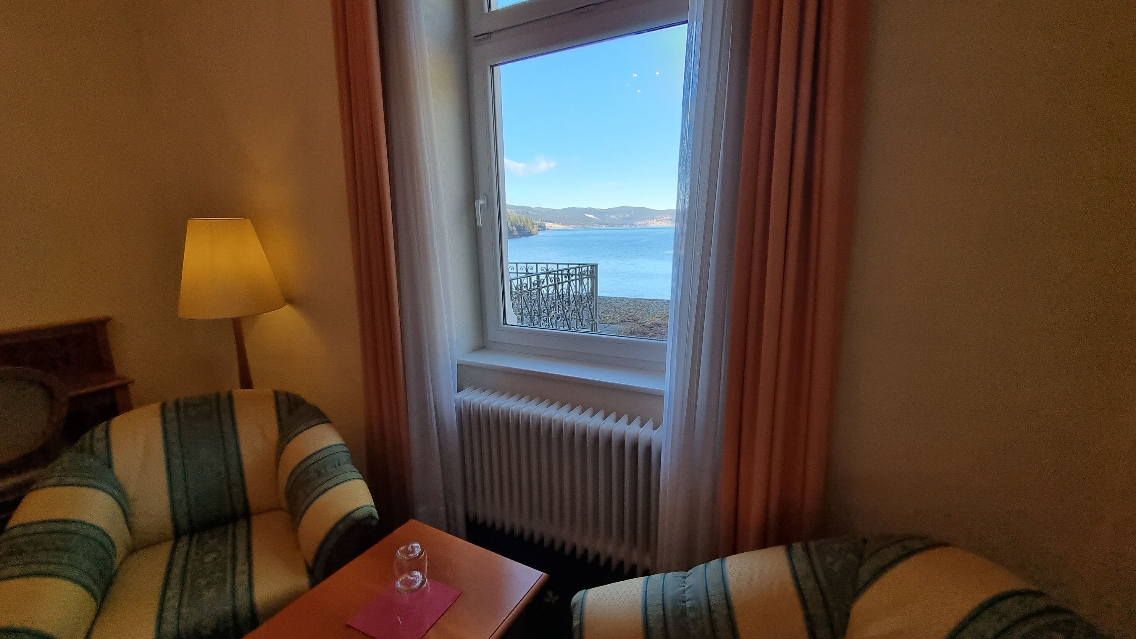 Une pièce avec vue sur un lac, visible par une fenêtre, comprenant un radiateur, une lampe et des fauteuils à rayures.