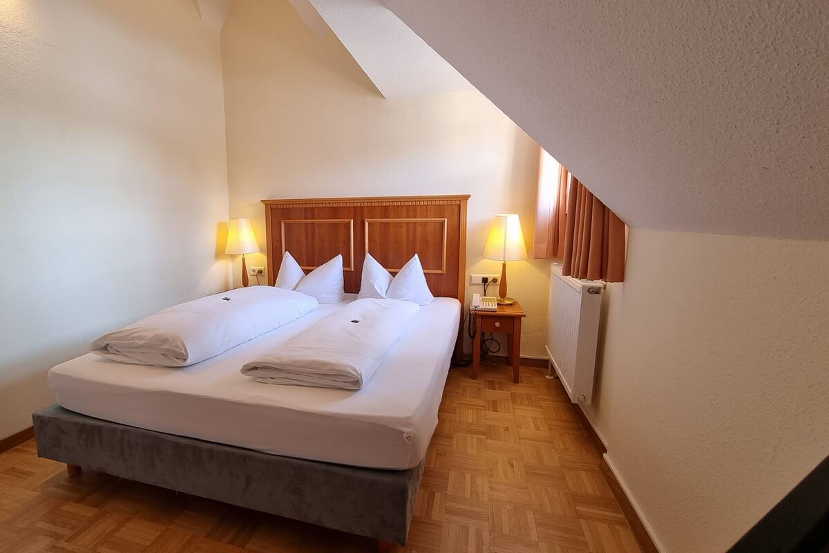 Een net opgemaakt tweepersoonsbed met twee kussens, een houten hoofdbord, twee bedlampjes, een klein nachtkastje met lamp en een radiator.