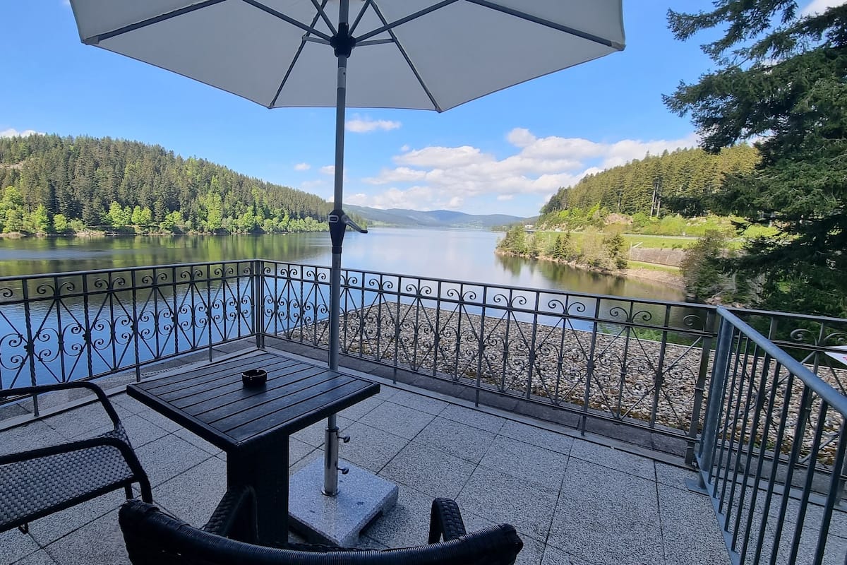 Een terras met een tafel en stoelen, met uitzicht op een rustig meer en beboste heuvels.