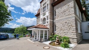 A stone building with a tiled roof, a covered entrance, and a small garden with a rock sculpture. - Seehotel Hubertus Schluchsee (Schluchsee)