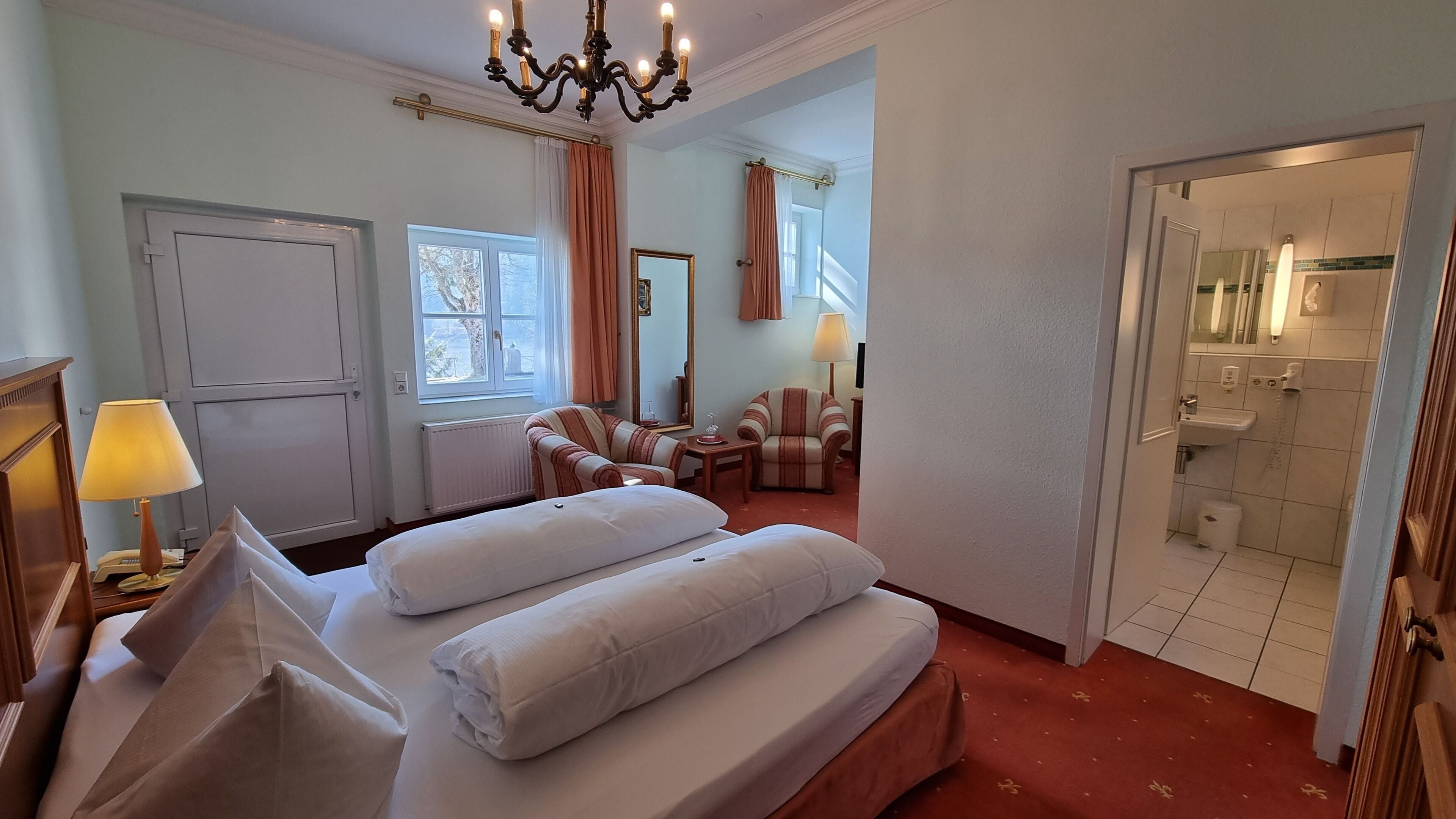 A hotel room with a bed, two armchairs, a small table, a mirror, and a bathroom visible through an open door.