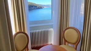 A room with a large window overlooking a body of water, two upholstered chairs, a round wooden table, and a radiator below the window. - Seehotel Hubertus Schluchsee (Schluchsee)