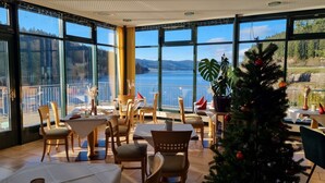A dining area with tables and chairs, large windows with a view of a lake and mountains, and a Christmas tree. - Seehotel Hubertus Schluchsee (Schluchsee)