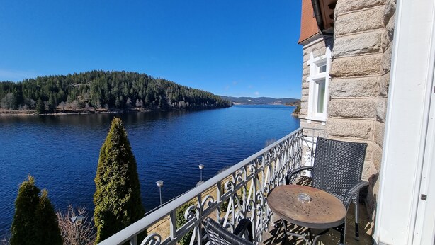 A balcony with a view of a lake, a stone building, and a metal railing. - Seehotel Hubertus Schluchsee (Schluchsee)