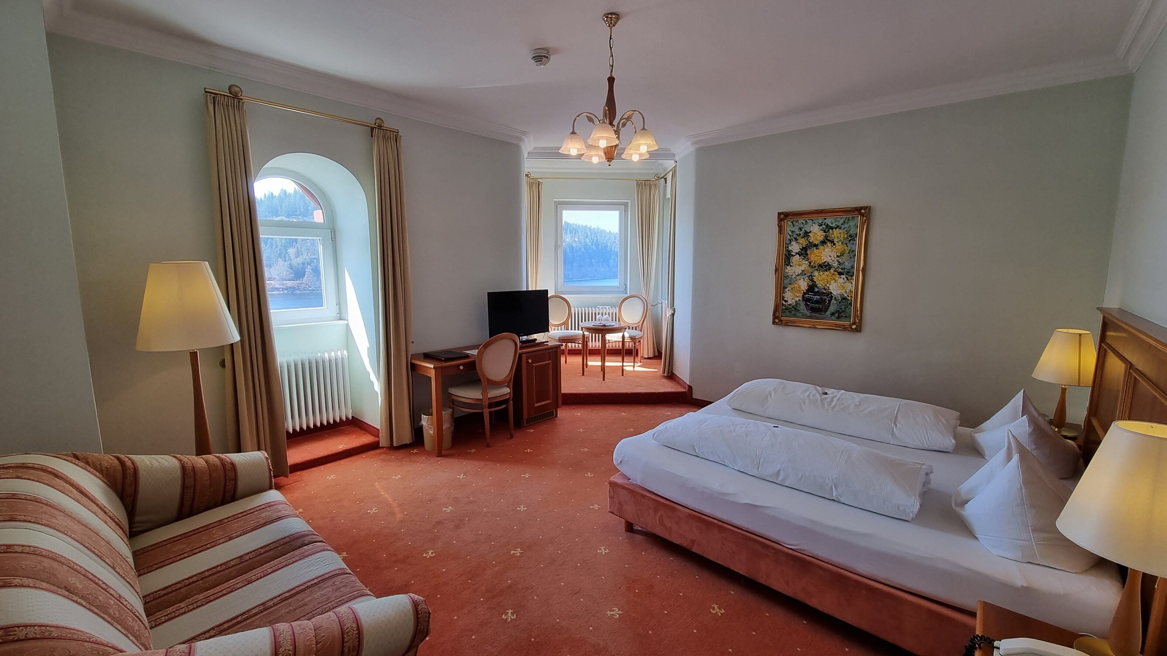 A hotel room with a bed, a desk with a chair, a television, and a painting on the wall.