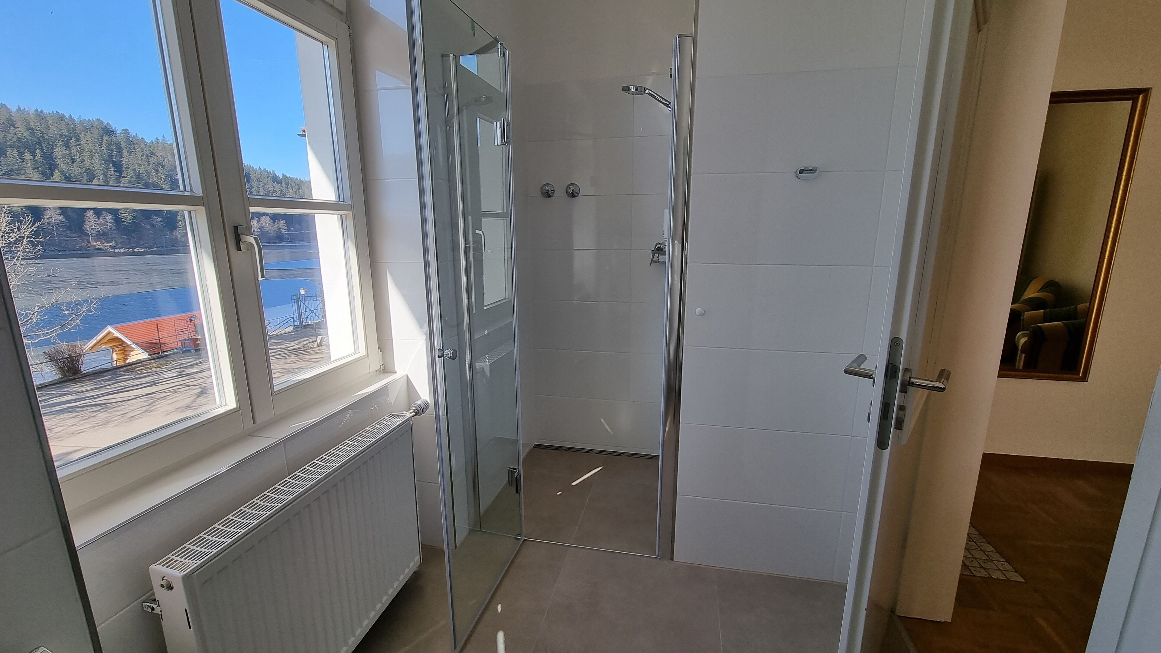 A bathroom with a glass shower door, a window with a view of a lake and buildings, and a radiator.