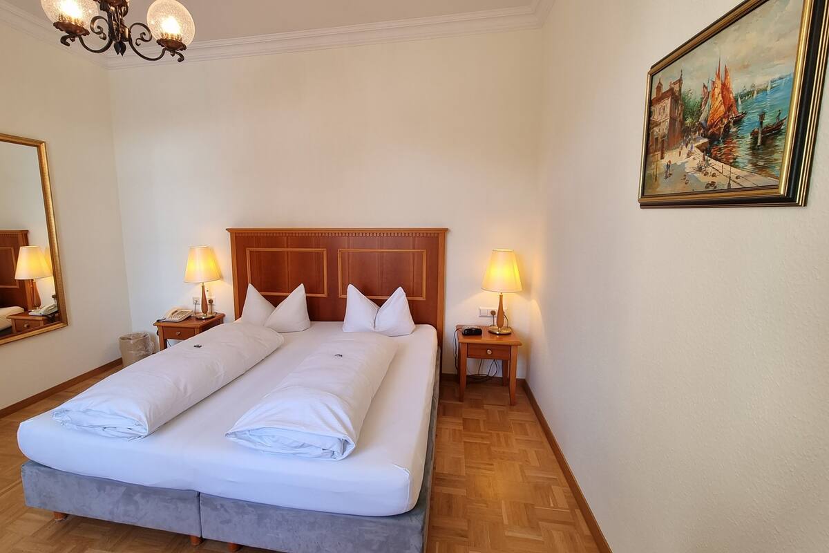 Een hotelkamer met een tweepersoonsbed, twee nachtkastjes met lampen, een houten hoofdeinde, een grote spiegel en een schilderij aan de muur.