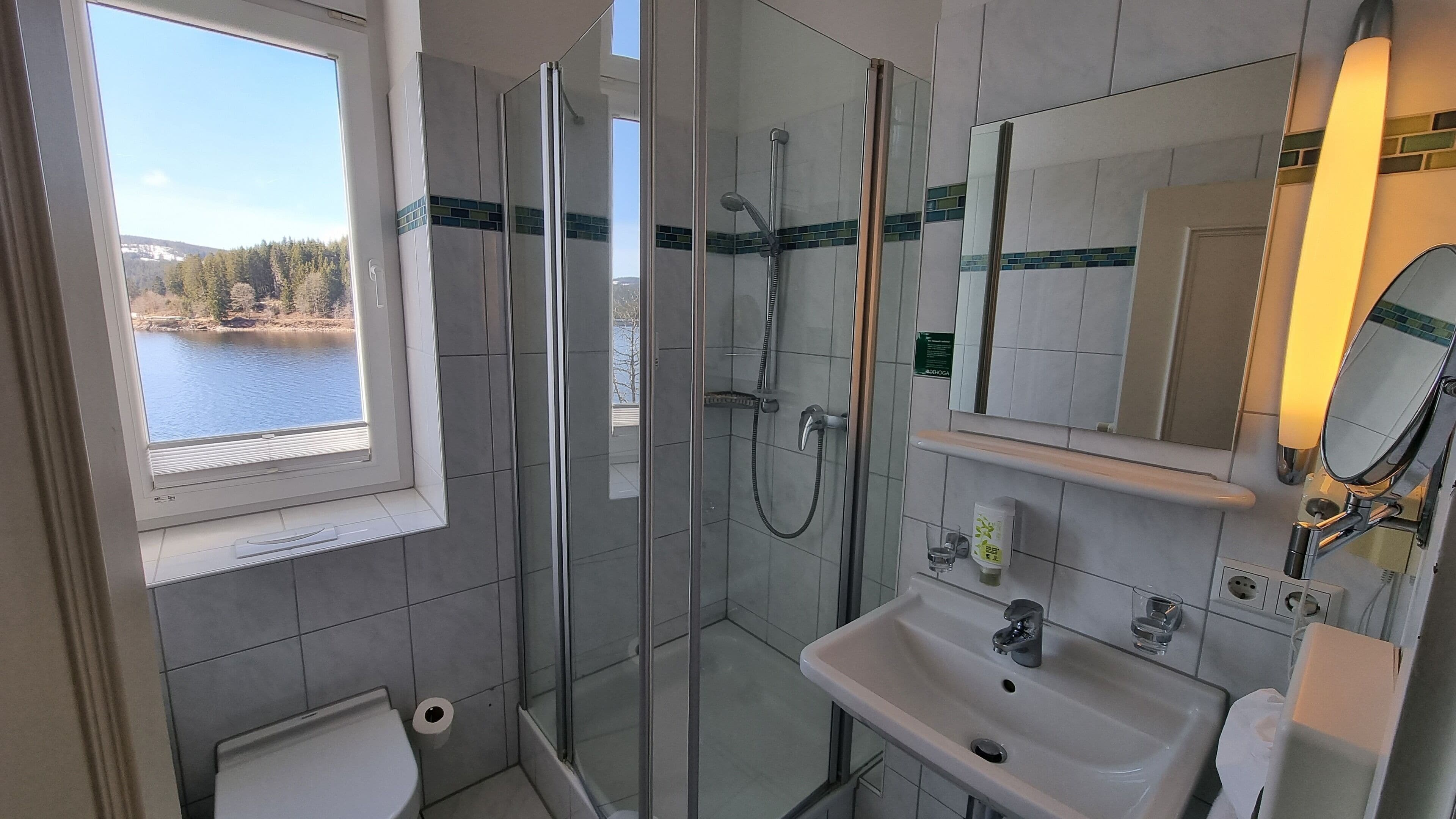 Un baño moderno con ducha envuelta en vidrio, lavamanos y un espejo grande. Una ventana ofrece vistas a un lago.