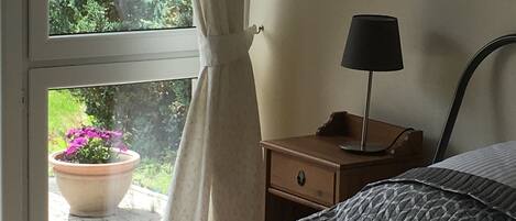 Un dormitorio con cama, mesita de noche con lámpara, ventana con cortinas y vistas al verdor exterior.