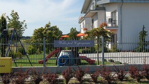 A gated playground area with a slide and swings, a sign reading "Willa pod Cyprysami," and a building with balconies and flowers. - Willa Pod Cyprysami (Ustka)