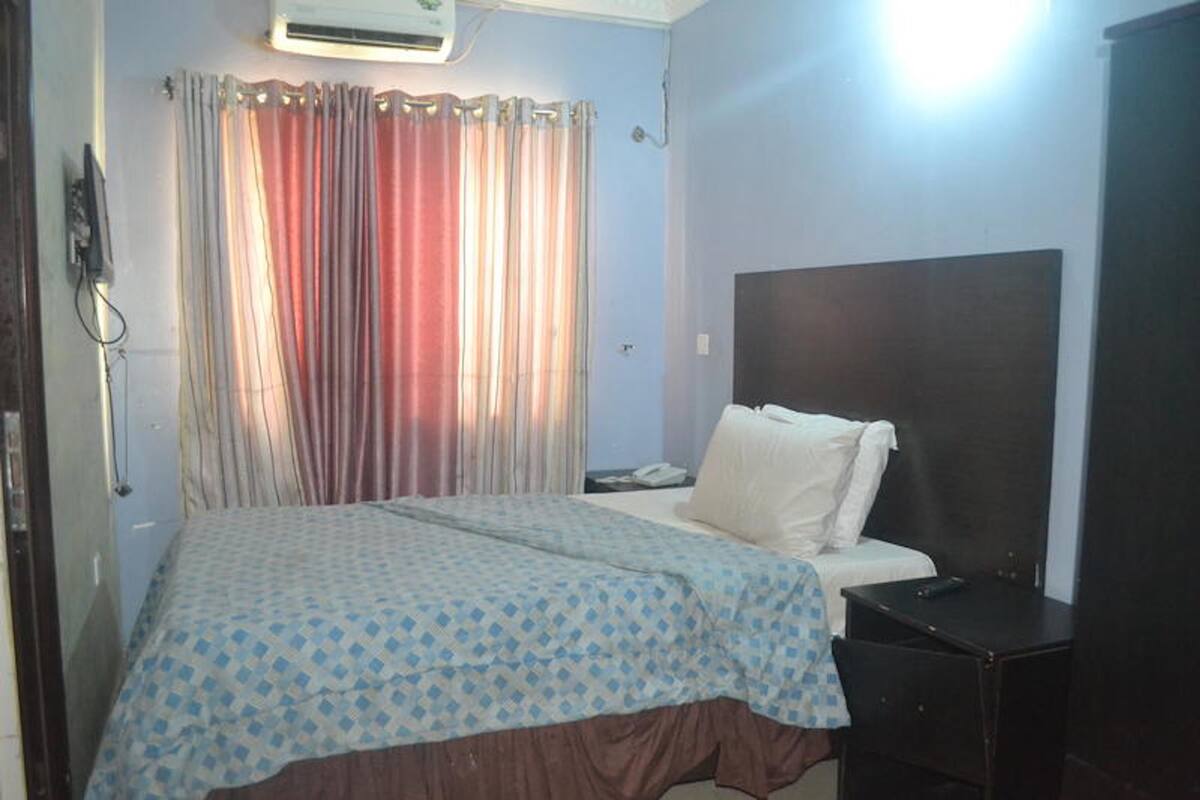 A bedroom with a bed, a bedside table, a nightstand, a curtain, and an air conditioning unit.