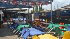 An outdoor seating area with colorful plastic chairs and tables, a shaded area with a sign, and a building with a signboard in the background. - Dan Morgan Hotel & Suites (Lagos)