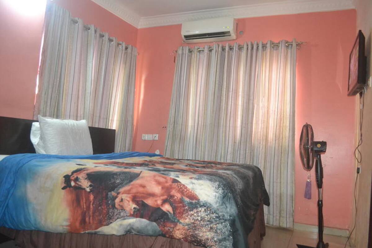 A bedroom with a bed, curtains, an air conditioner, a fan, and a television on the wall.