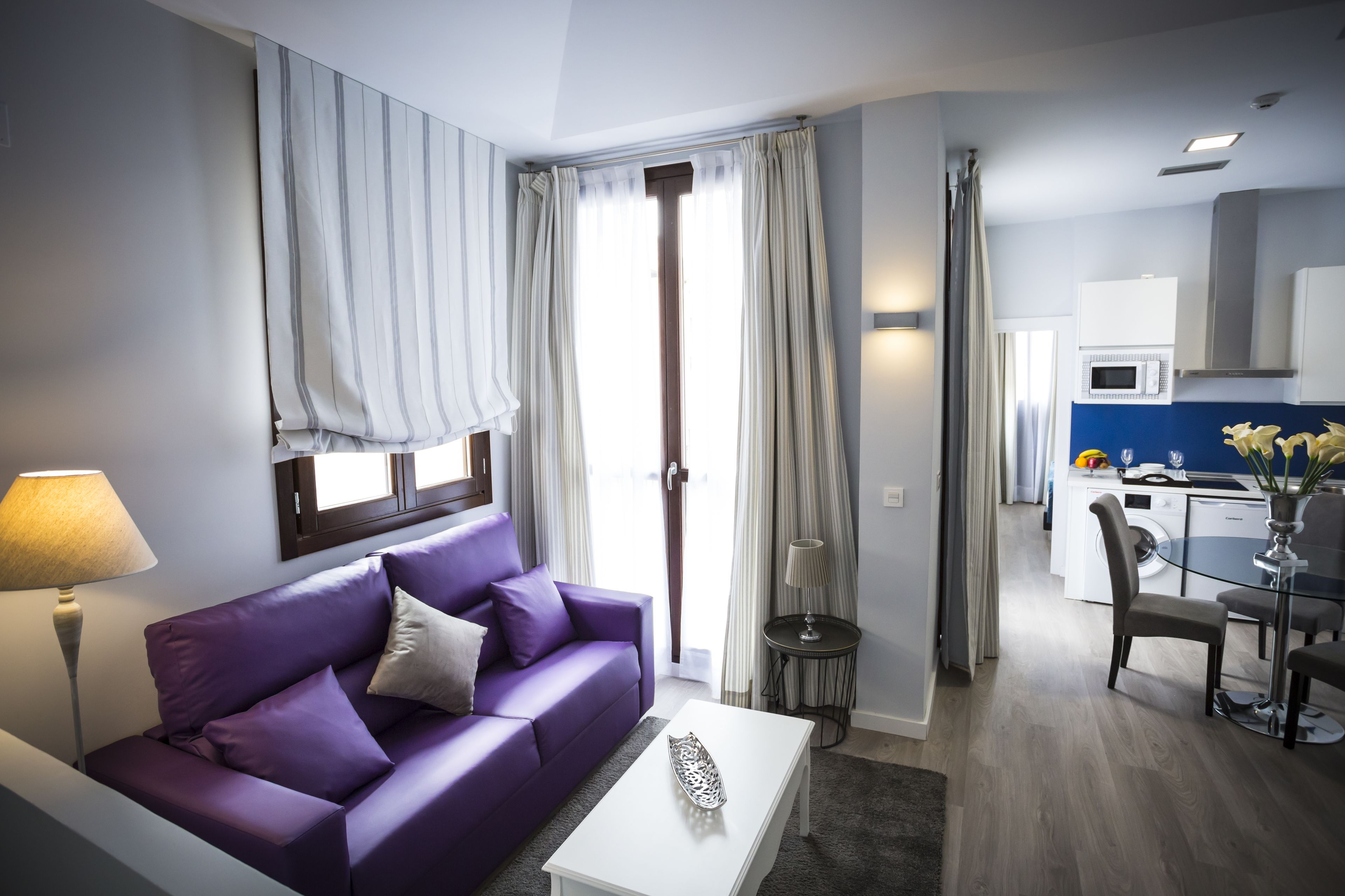 Una sala de estar moderna con un sofá morado, una mesa de centro blanca y un comedor con una mesa y sillas de vidrio.