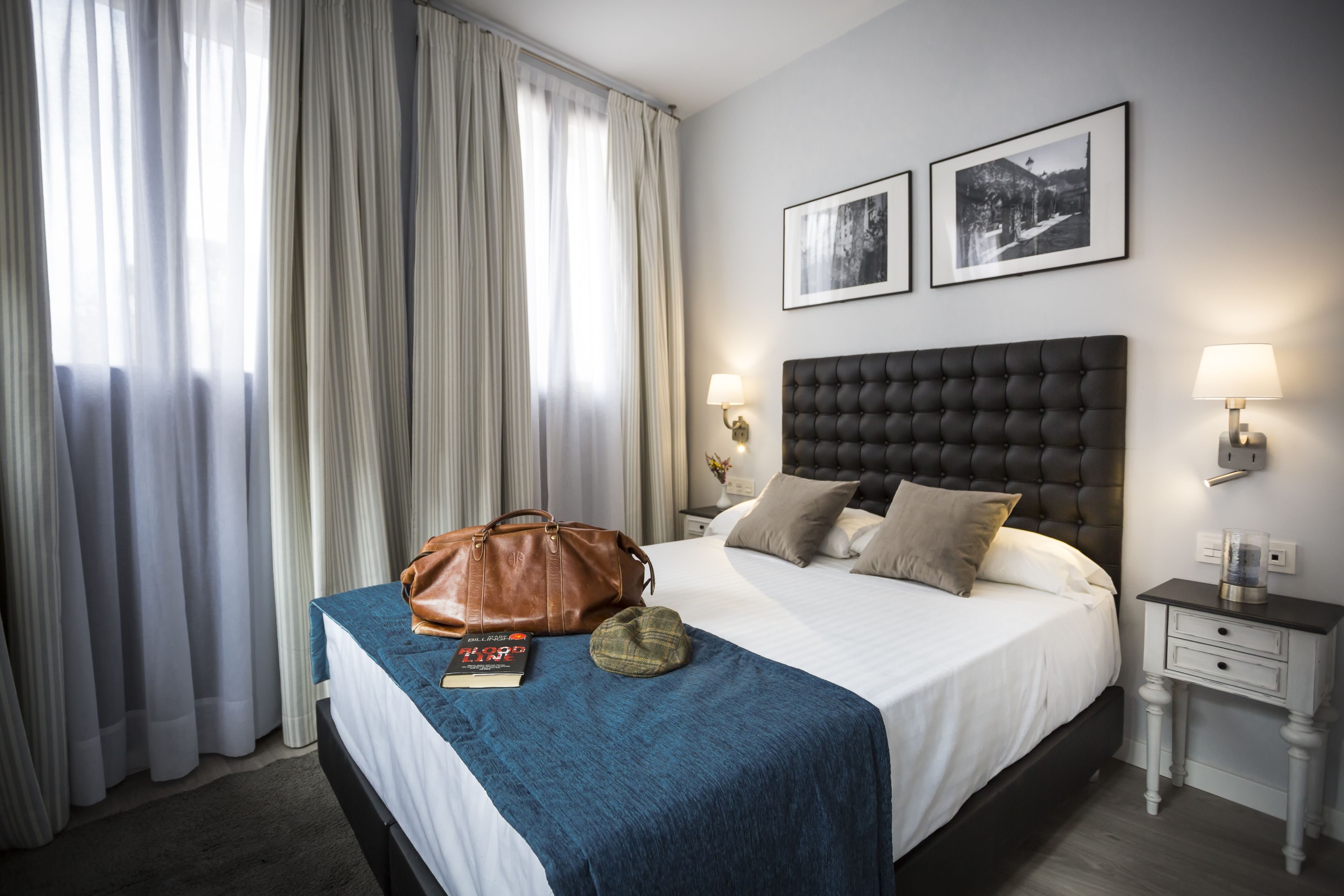 A neatly made bed with a blue blanket, a brown leather bag, and a book on the bed. There are two framed pictures on the wall, a lamp on the nightstand, and a window with curtains.