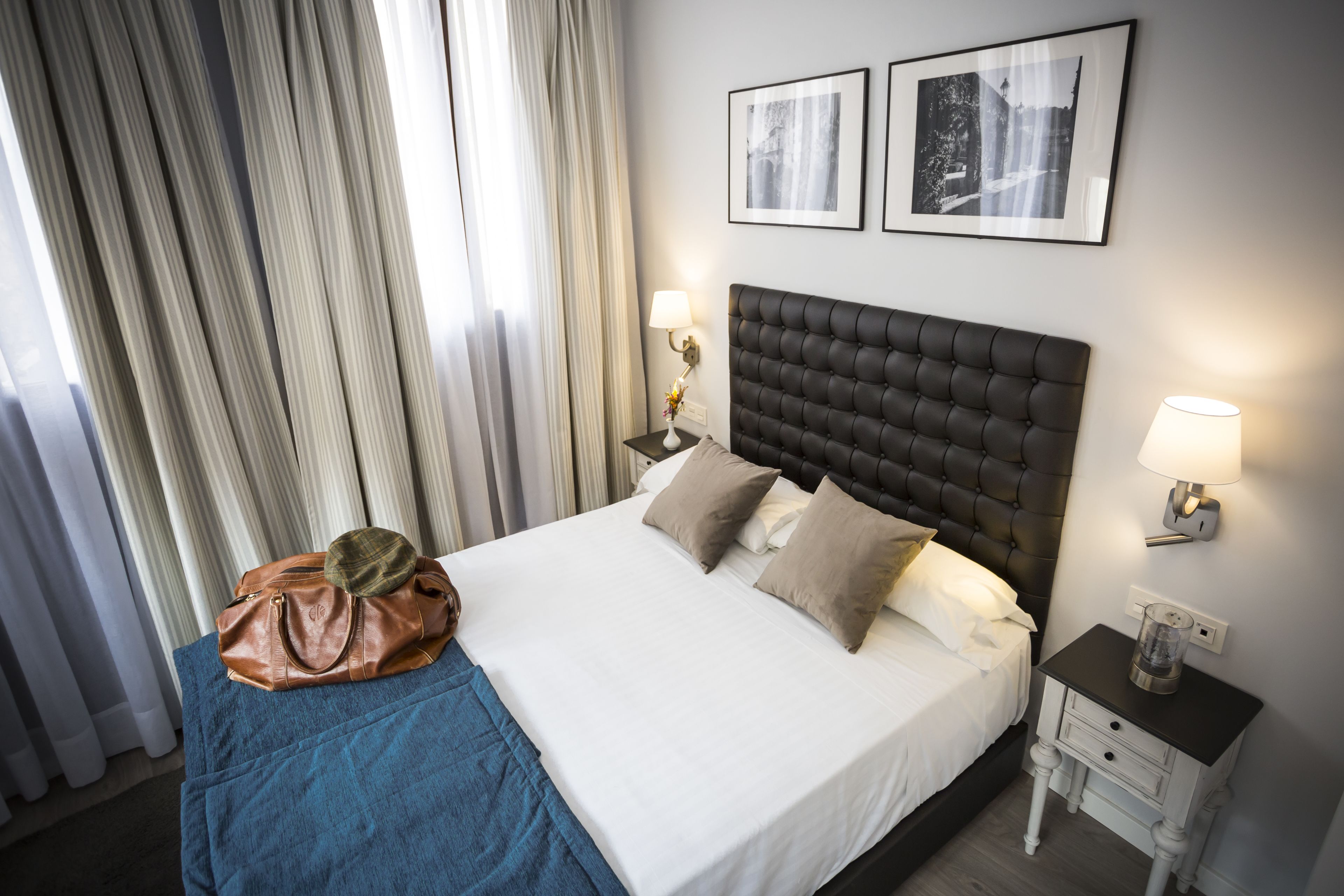A neatly made bed with a dark headboard, a brown leather bag, and a blue blanket on a wooden bed frame. There are two bedside tables with lamps and a wall-mounted picture frame.