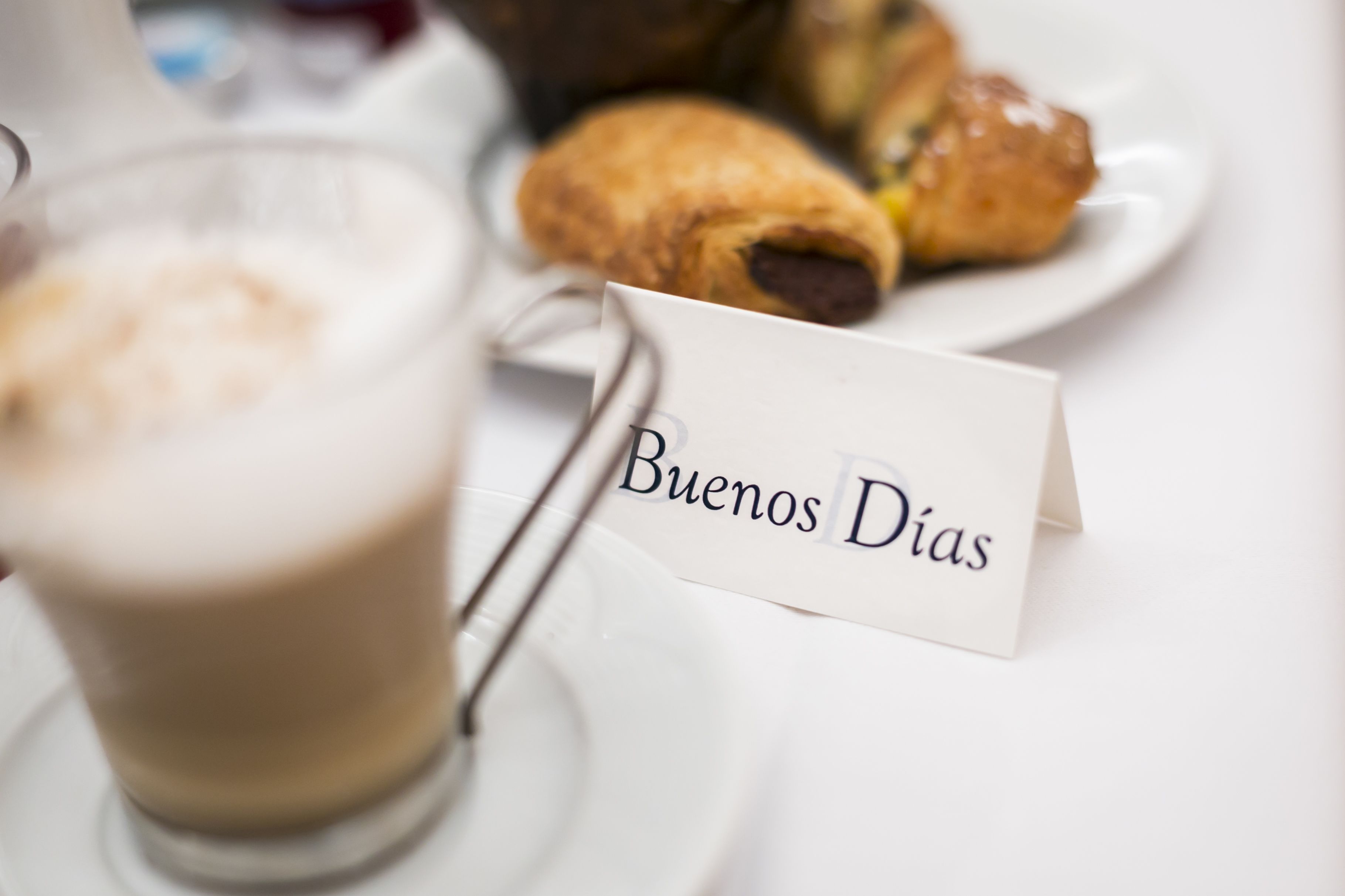 A table setting with a glass of coffee and a plate of pastries, and a "Buenos Dias" sign.