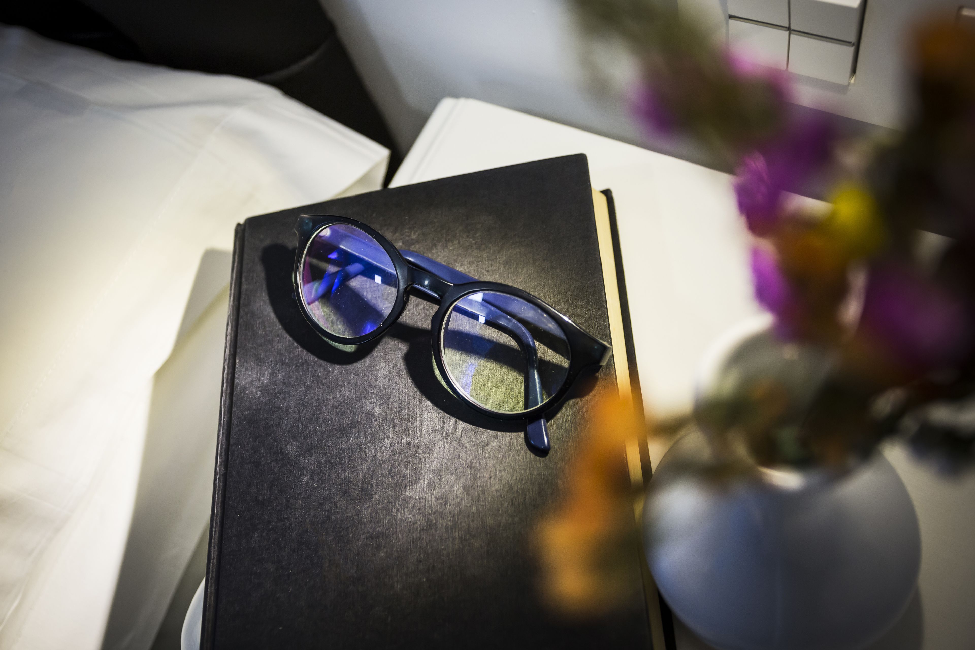 A pair of glasses on a black box, a white pillow, and a vase with flowers.