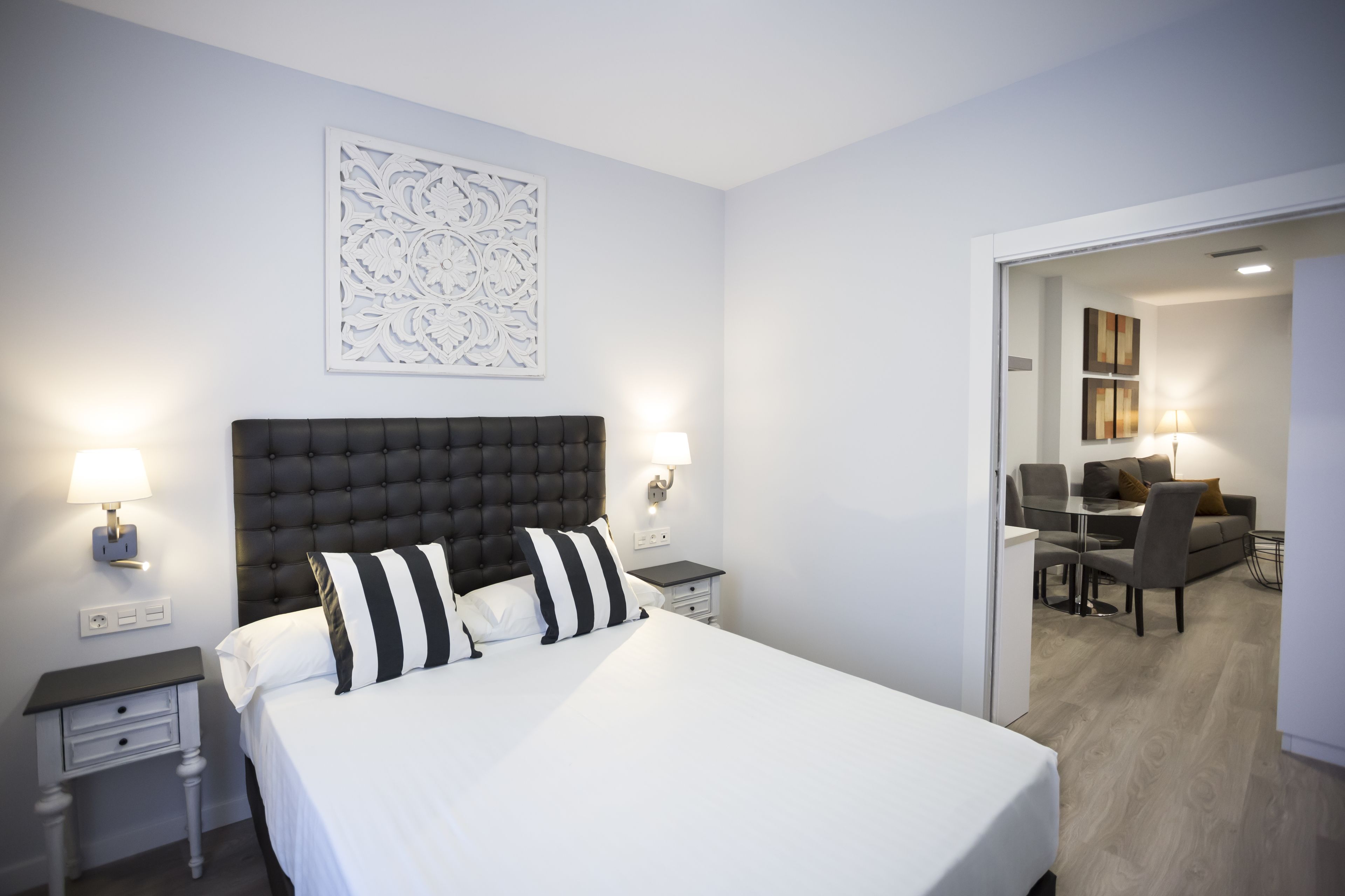 Ein modernes Hotelzimmer mit einem großen Bett, einem Nachttisch, einer Wandleuchte und einem Essbereich, der durch eine geöffnete Tür sichtbar ist.