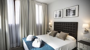 Un lit bien fait, recouvert d’une couverture bleue, avec deux oreillers et deux serviettes blanches posées sur une tête de lit sombre. Deux tableaux encadrés ornent le mur, ainsi qu’une table de chevet avec une lampe et une fenêtre avec des rideaux.