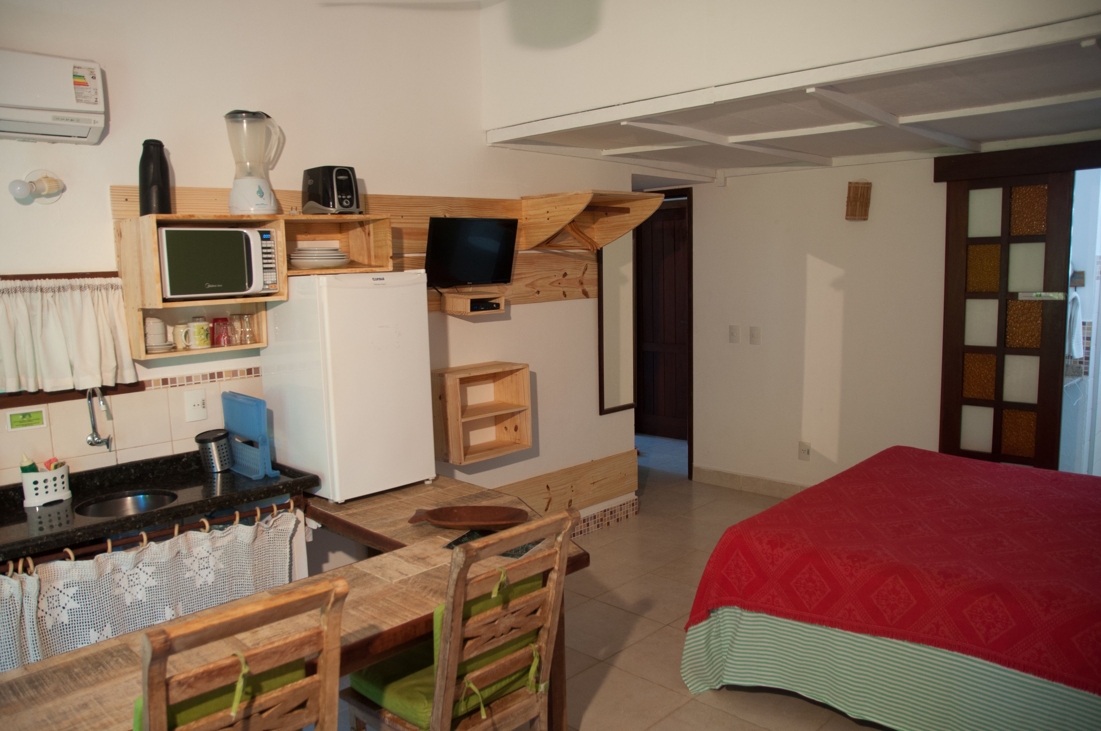 A compact kitchen with a microwave, stove, and sink. There is a small wooden table with chairs and a red bed in the adjacent room.