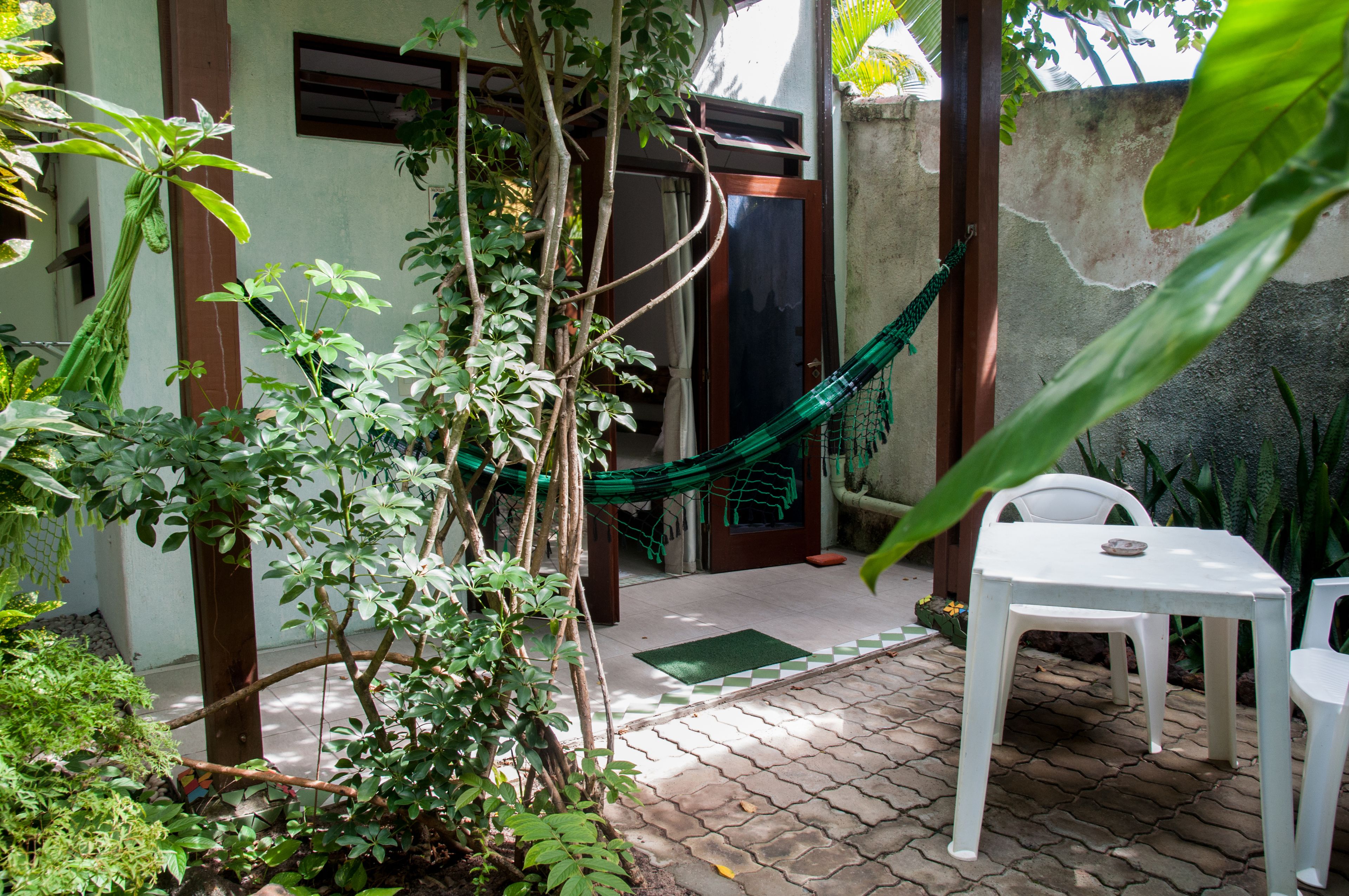 Un patio con mesa y sillas blancas, rodeado de follaje y una hamaca.