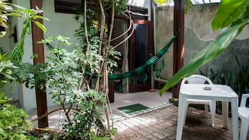 Un patio con mesa y sillas blancas, rodeado de follaje y una hamaca.