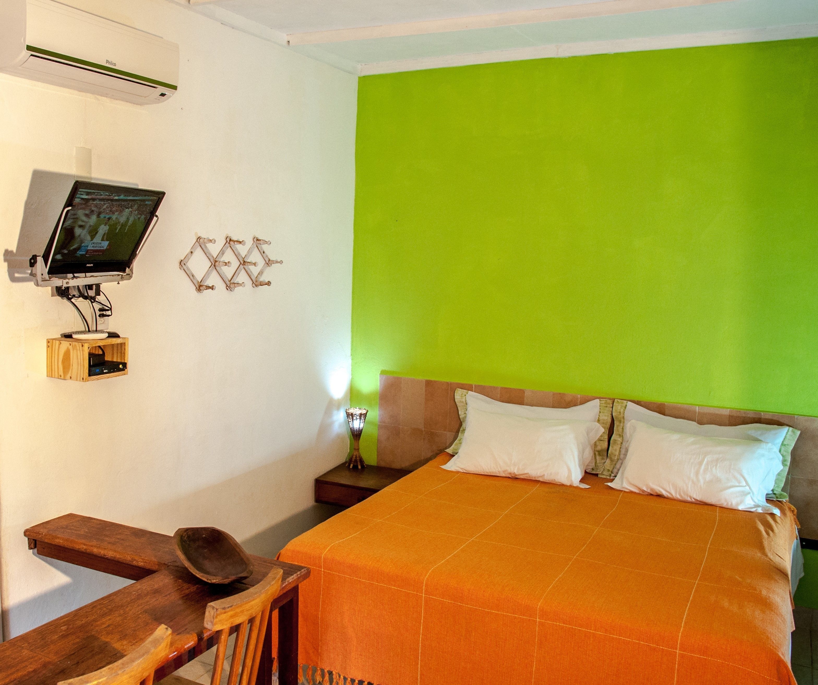 Une chambre avec un mur vert, un lit recouvert d’une couverture orange, une table de chevet en bois, un bureau avec un écran d’ordinateur et un climatiseur fixé au mur.