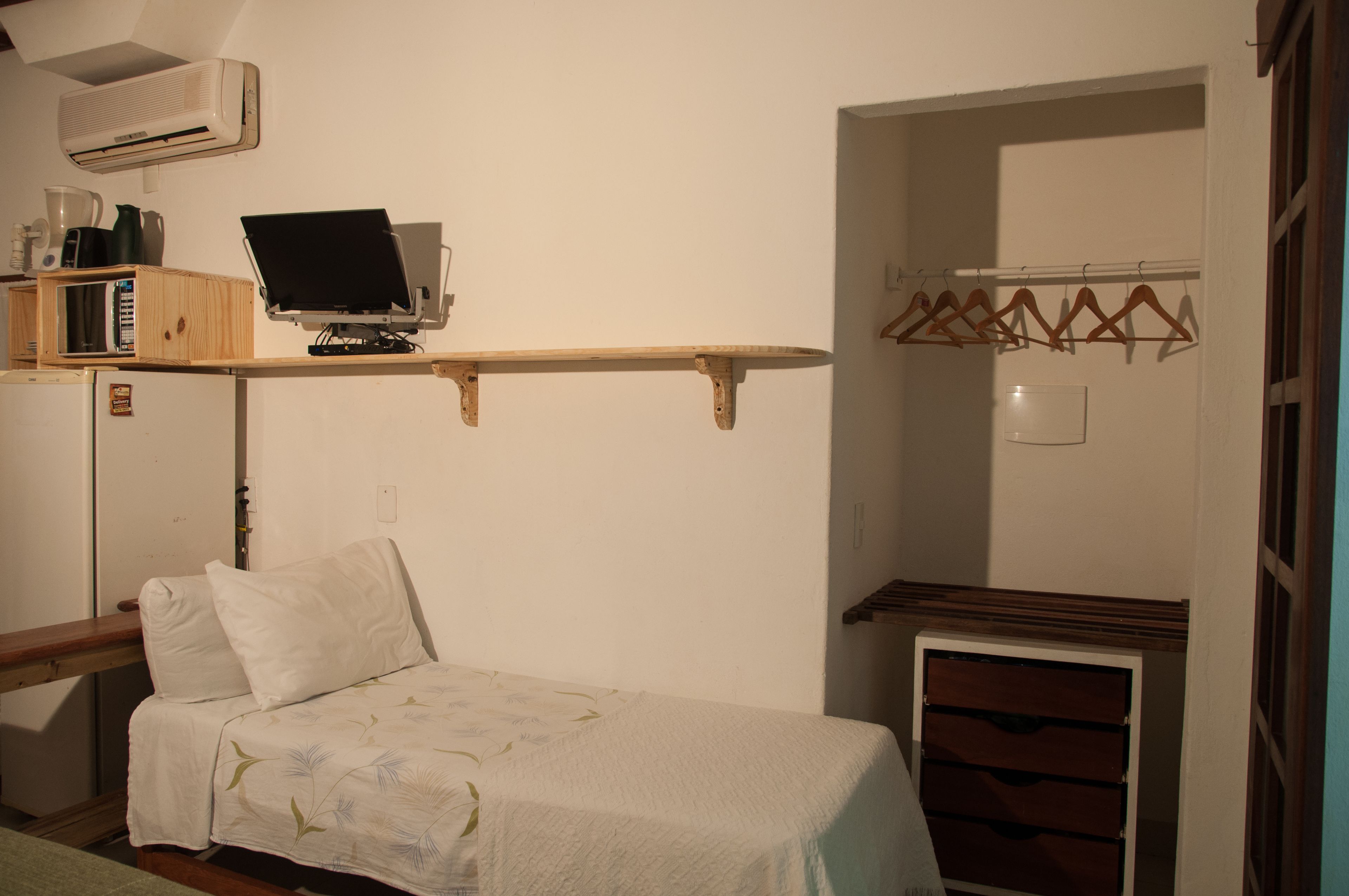 Pequeña habitación con cama, escritorio con ordenador, estante con libros y hervidor, y perchero con perchas.