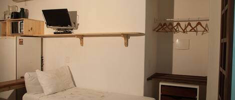 Pequeña habitación con cama, escritorio con ordenador, estante con libros y hervidor, y perchero con perchas.